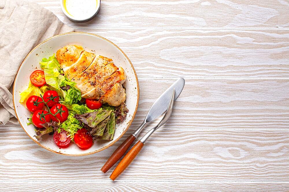 Healthy green vegetable salad with grilled chicken breast fillet on ceramic plate with olive oil on the side on white wooden kitchen table top view flat lay, diet food concept with space for text