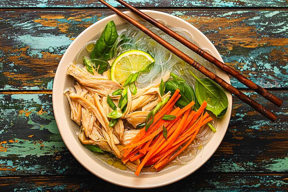 Asian soup with rice noodles, chicken and vegetables in ceramic bowl served with spoon and chopsticks on rustic wooden background from above, Chinese or Thai cuisine