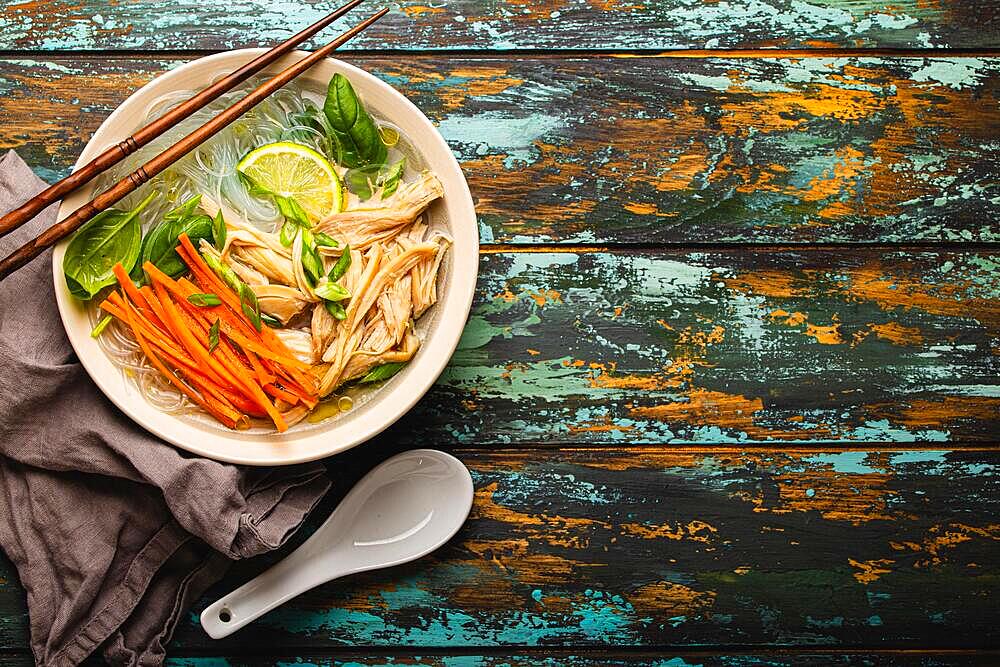 Asian soup with rice noodles, chicken and vegetables in ceramic bowl served with spoon and chopsticks on rustic wooden background from above with space for text, Chinese or Thai cuisine