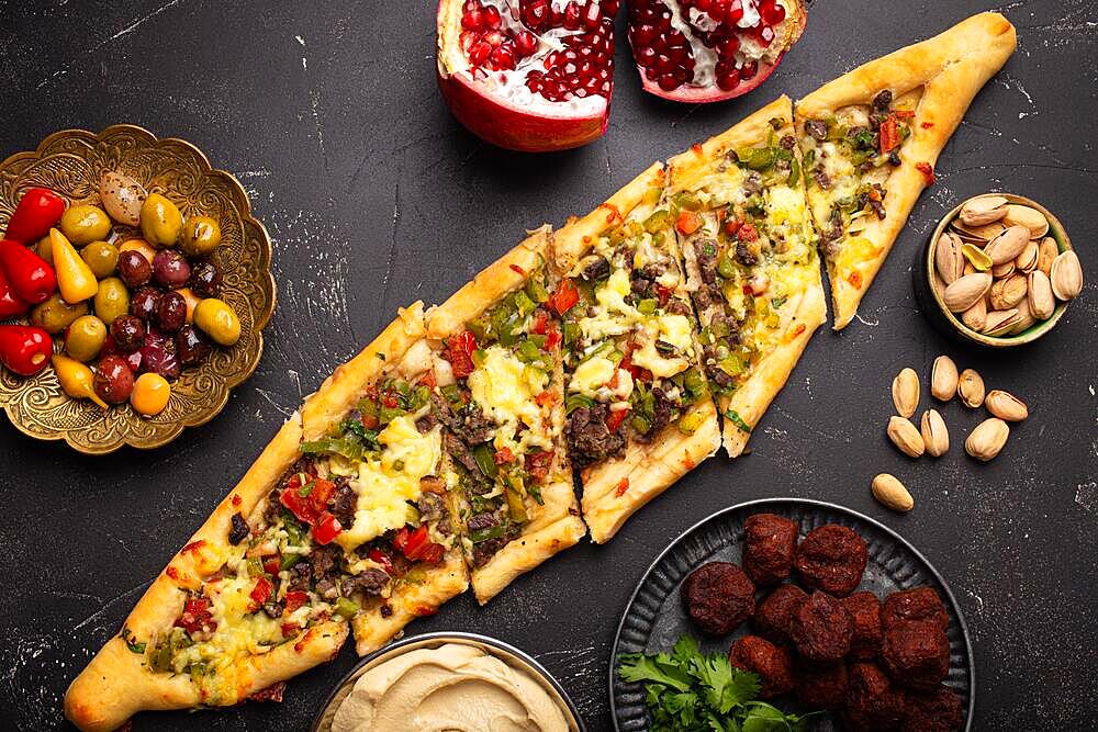 Traditional Turkish pizza pide freshly baked with ground beef and vegetables served on black stone background with assorted Middle Eastern meze from above, cuisine of Turkey