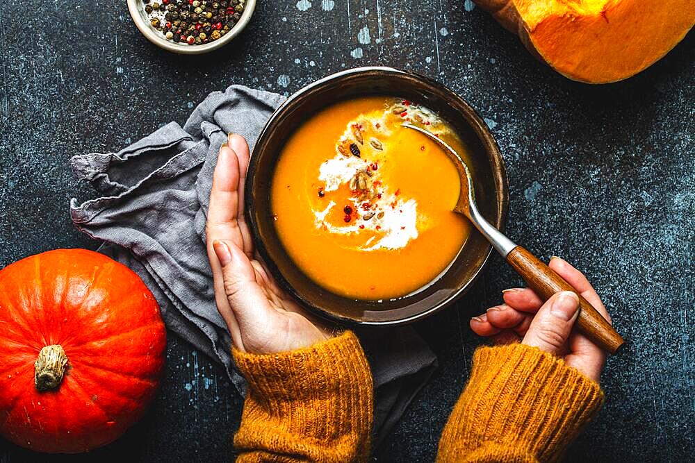 Woman in yellow knitted sweater eating a pumpkin cream soup on dark stone background with spoon decorated with cut fresh pumpkin, top view. Autumn cozy dinner concept