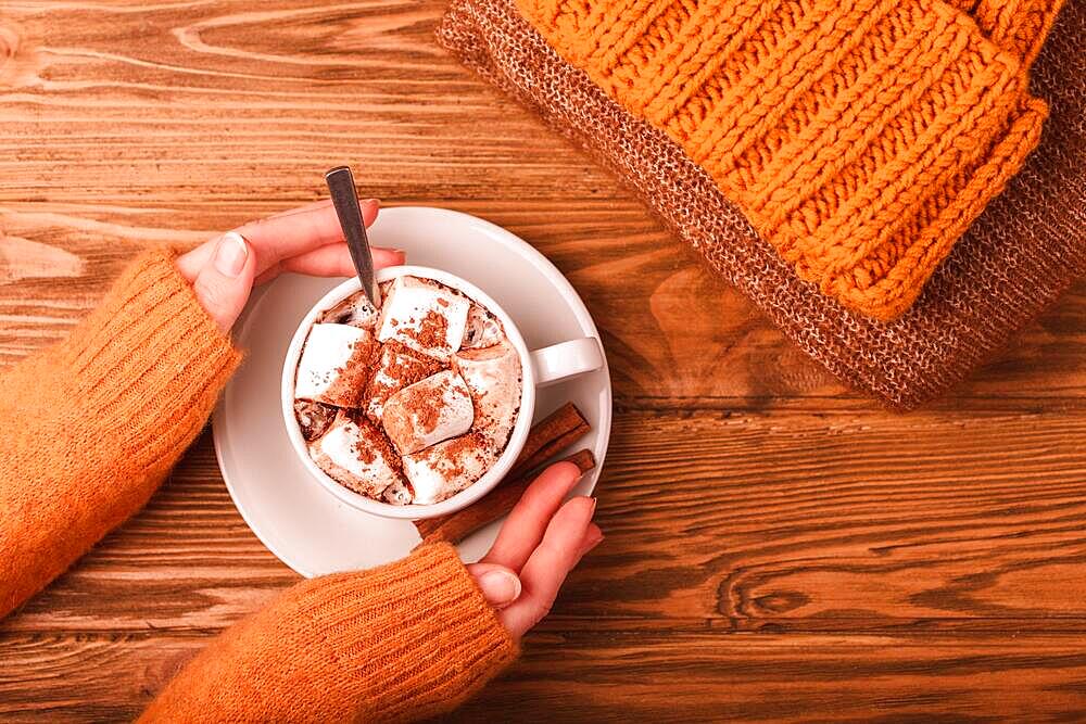 Cozy winter flat lay, female hands holding cup with hot chocolate or cocoa with marshmallow and pile of warm knitted clothes on wooden rustic background. Comfy and soft winter or autumn concept