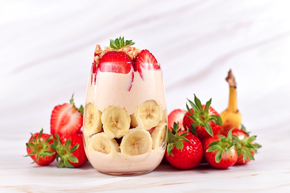 Layered banana and strawberry fruit summer dessert with yogurt and granola in glass surrounded by fresh ingredients