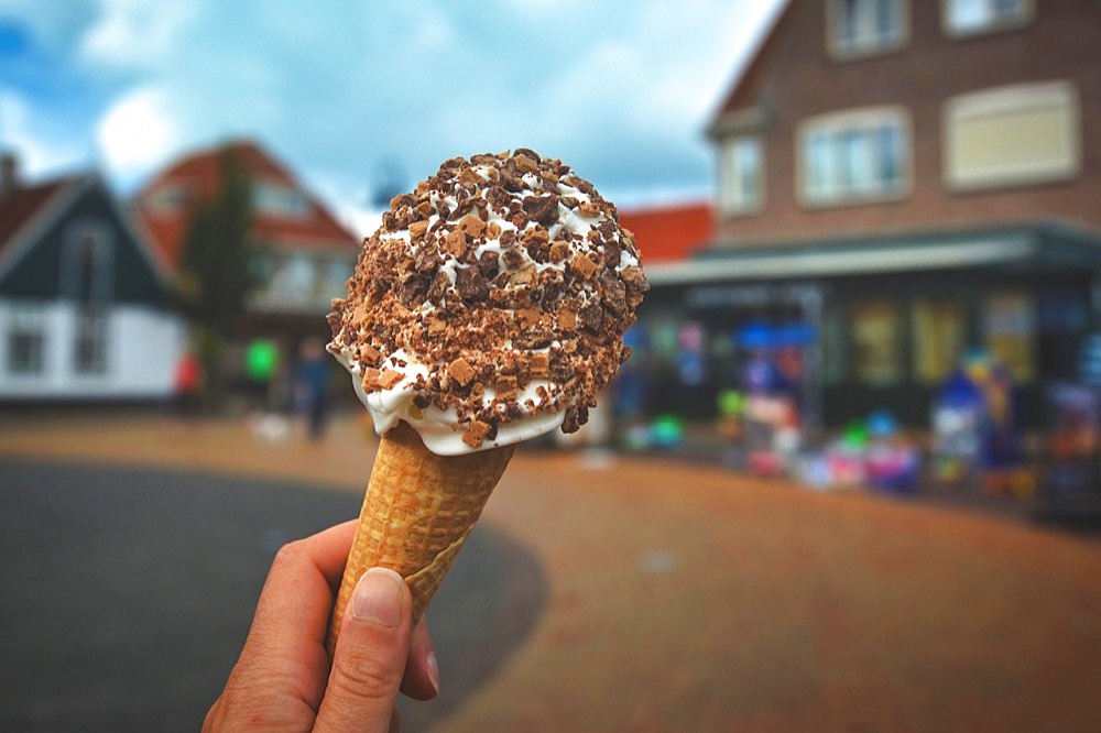 Close up of hand holding cone with white soft serve ice cream covered in chocolate sprinkles with blurry city scene in background