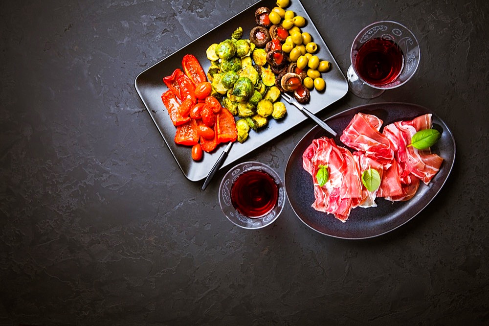 Prosciutto with different appetizers with glass of red wine on black background. Over roasted vegetables, healthy snack