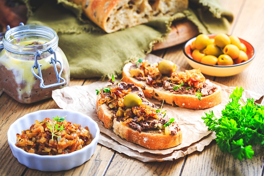 Fresh homemade chicken liver pate on ciabatta bread with roasted onions and olives on wooden table