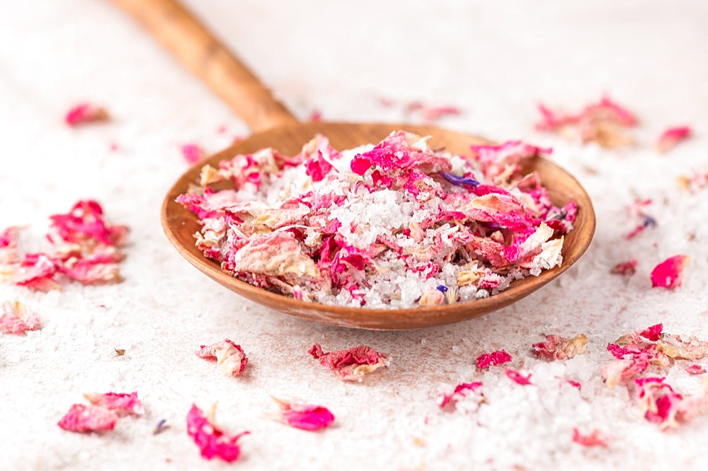 See salt wih with dried flower petals in wooden scoop on pink marble table