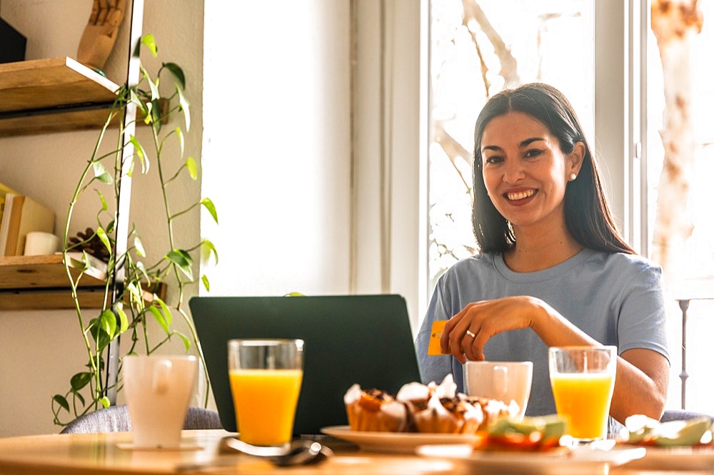 Woman shopping online with computer while having breakfast in the morning by the window with orange juice