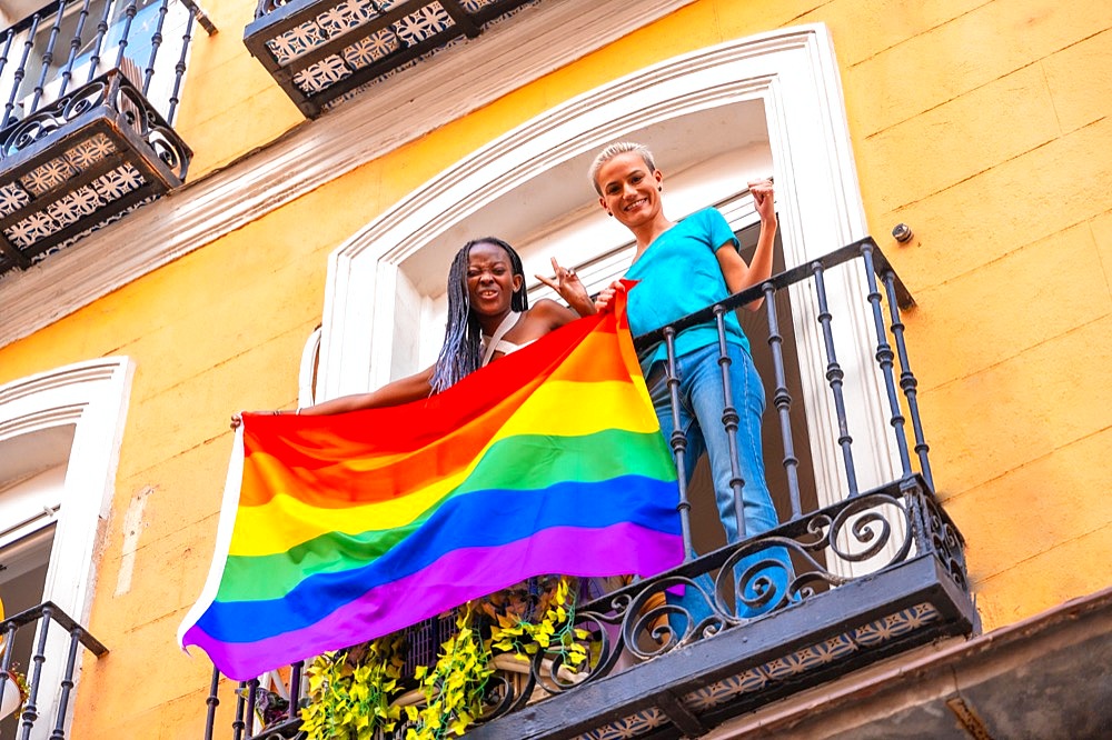 Lesbian girl couples with lgbt flag on balcony at home party, waving smiling, LGBT pride