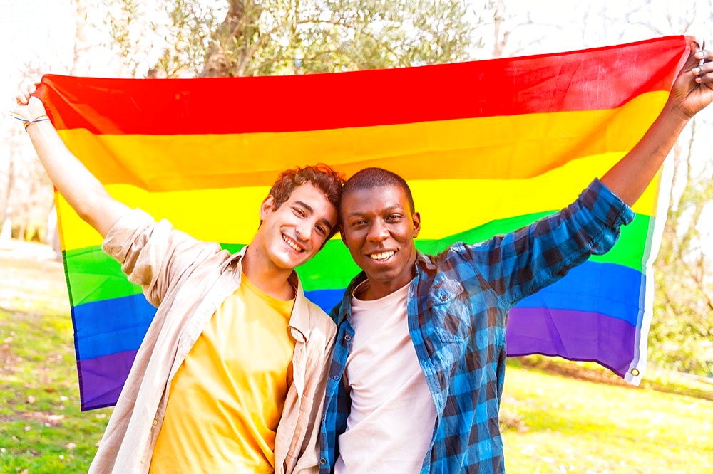 Portrait of multiethnic gay male couple holding a rainbow lgbt symbol flag at sunset