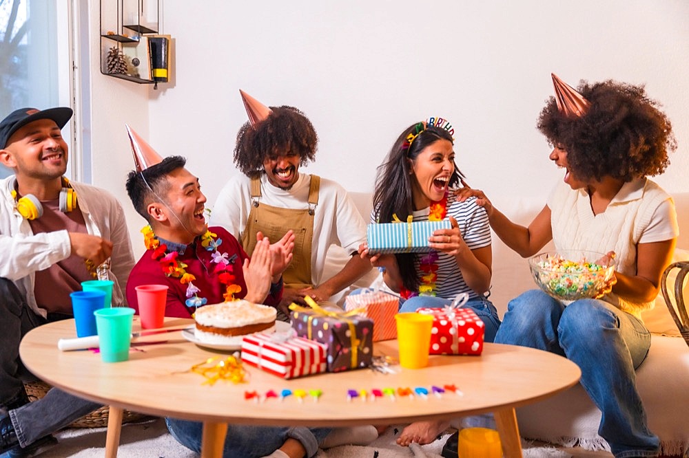 Group of friends at a birthday party on the sofa at home smiling delivering the gifts, surprising the birthday girl