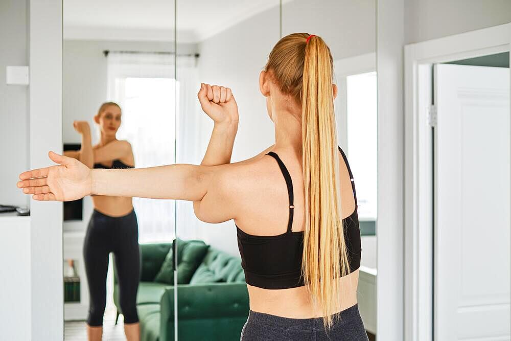 Young woman in sportswear stretches arms in her living room in front of a mirror, back view