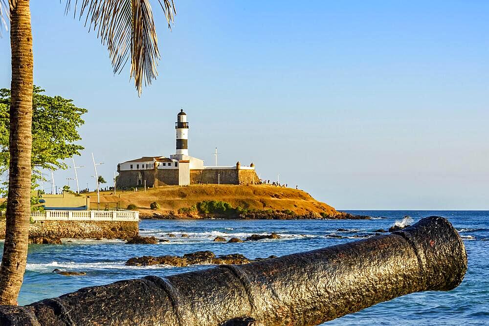 Old cannon and the Barra Lighthouse (Farol da Barra) one of the main historical buildings and tourist spot in the city of Salvador in Bahia surrounded by the sea during the late afternoon., Brasil