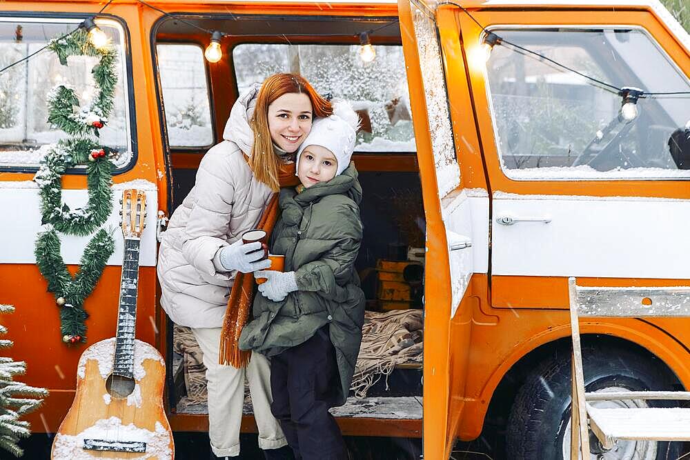 Lovely photo of young happy mother with little daughter in warm clothes hugging and drinking cocoa near orange minivan outdoors, family enjoying frosty winter weather. Winter holidays concept