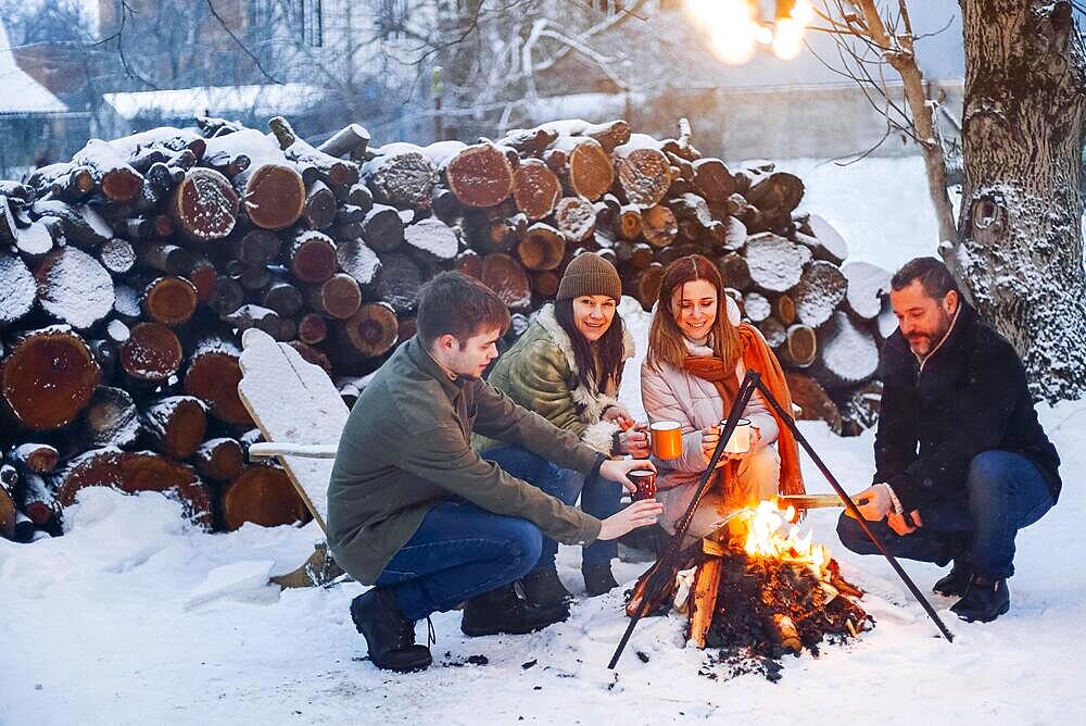 Group of friends gathering around bonfire in backyard, drinking tea and warming hands. Two happy couples relaxing and enjoying winter season while sitting around fire. Outdoor winter entertaining