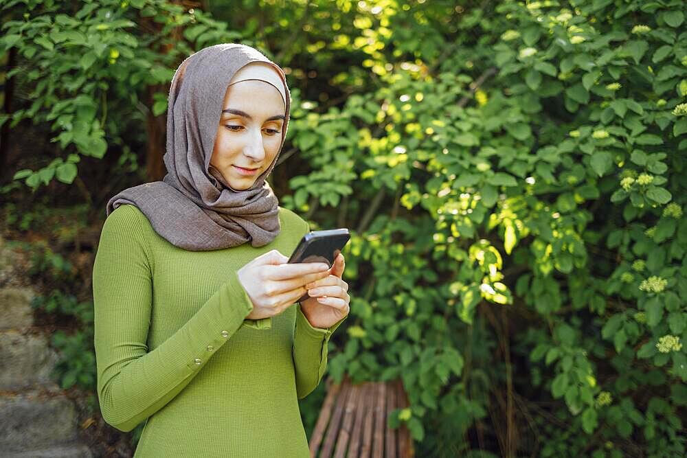 Modern Muslim girl student wearing hijab in the park, holding a phone in her hand and smiling