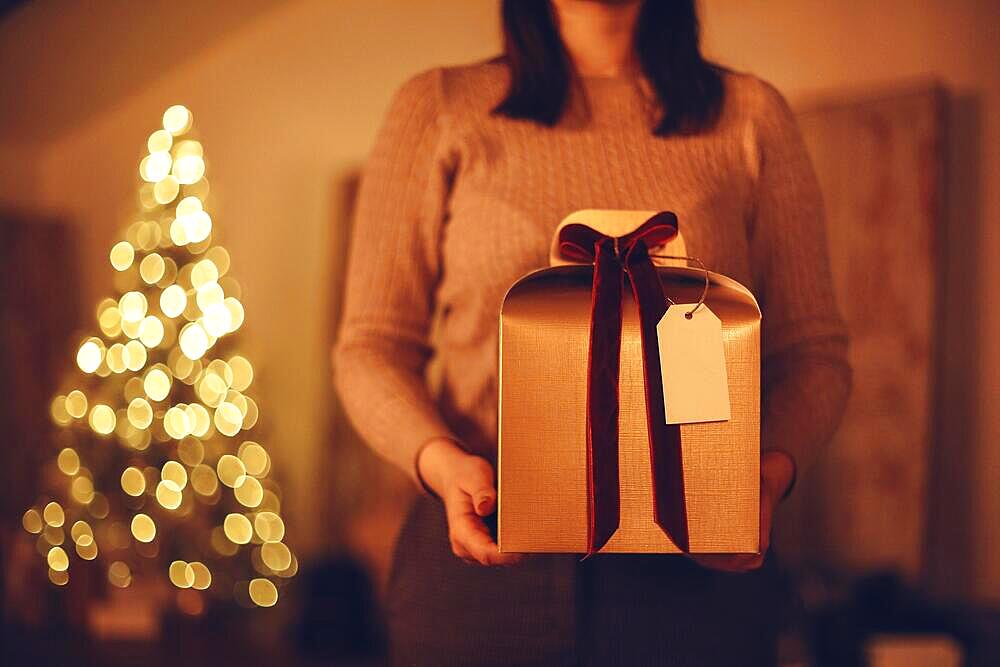 Christmas gift. Unrecognizable woman holding big golden xmas present box with red bow while standing against blurred xmas tree background, selective focus of holiday gift in female hands