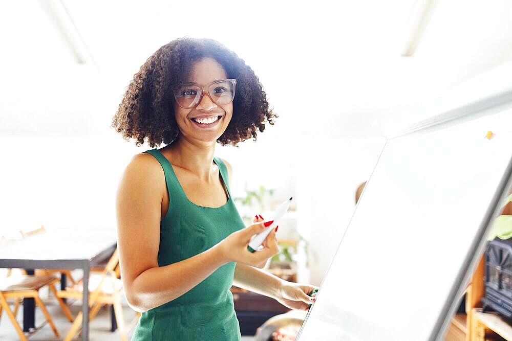 Beautiful African American girl with dark curly hair standing near board and giving presentation to colleagues in office. Young business woman looking on her coworkers while discussing new project