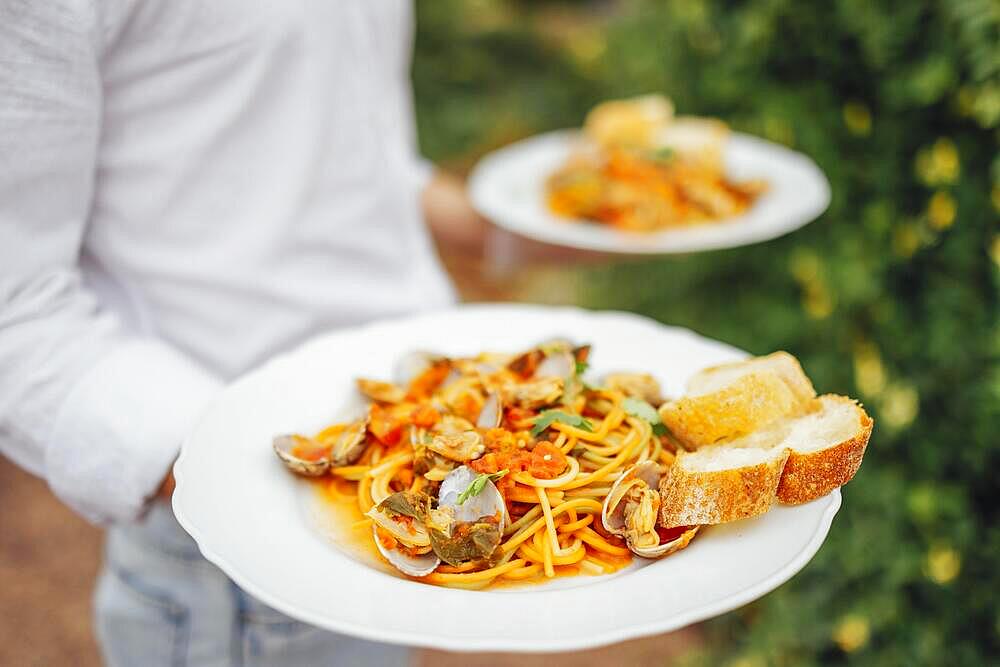 Seafood Spaghetti with seashells, prawns, squids hands of the waiter on ature background. Italian mediterranean food concept. Outdoors party