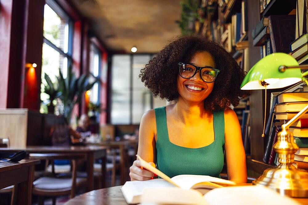 Happy African American female student studing in university library and looking at camera, wearing glasses by green lamp. Smiling teen girl in cozy library