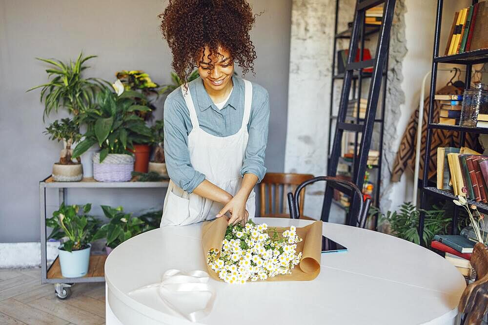 Tender shop assistant enjoying her job while serving bouquet for sale. Young florist in apron creating beautiful simple daisy bouquet in craft paper. Portrait of smiling girl with dark curly hair looking gladly at chamomile bouquet