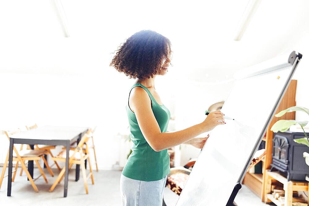 Beautiful African American girl with dark curly hair standing near board and giving presentation to colleagues in office. Young business woman looking on her coworkers while discussing new project