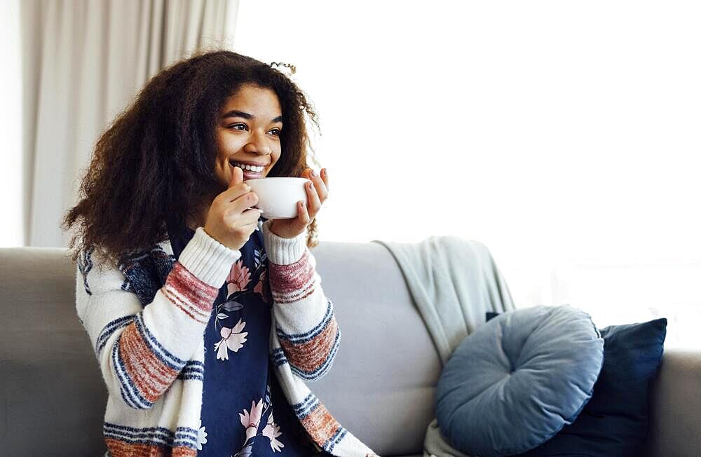 Cheerful smiling african american girl relaxing on couch with cup of coffee, resting at home, enjoying day off, wearing warm clothes