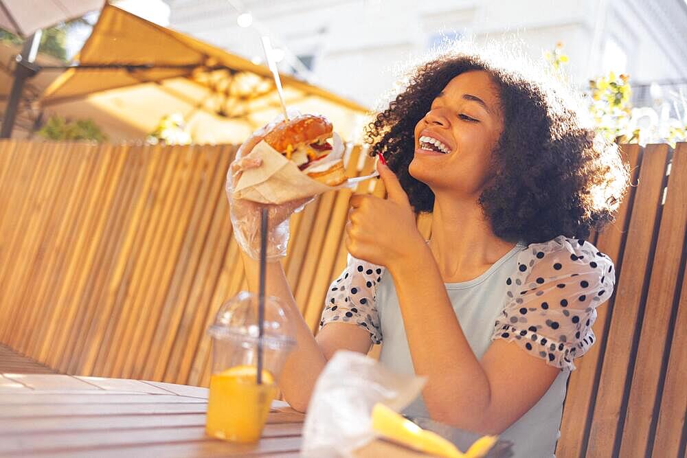 Young african american girl eating classic burger and drinking lemonade in the city. Outdoor woman wearing summer blouse with polka dots eatting delicious burger while sitting at terrace. Woman in blue tasting sandwich having joyful look admiring sunny w