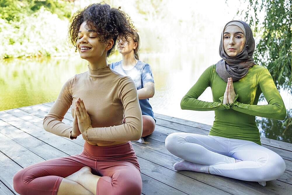 Group of teenages is engaged in fitness on pier by the lake or river. Young and positive girls go in for sports. Muslim in a hijab, african american and white females are doing stretching in the park