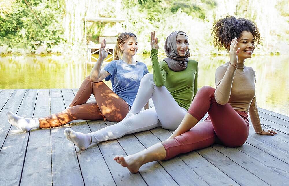 Group of teenages is engaged in fitness on pier by the lake or river. Young and positive girls go in for sports. Muslim in a hijab, african american and white females are doing stretching in the park