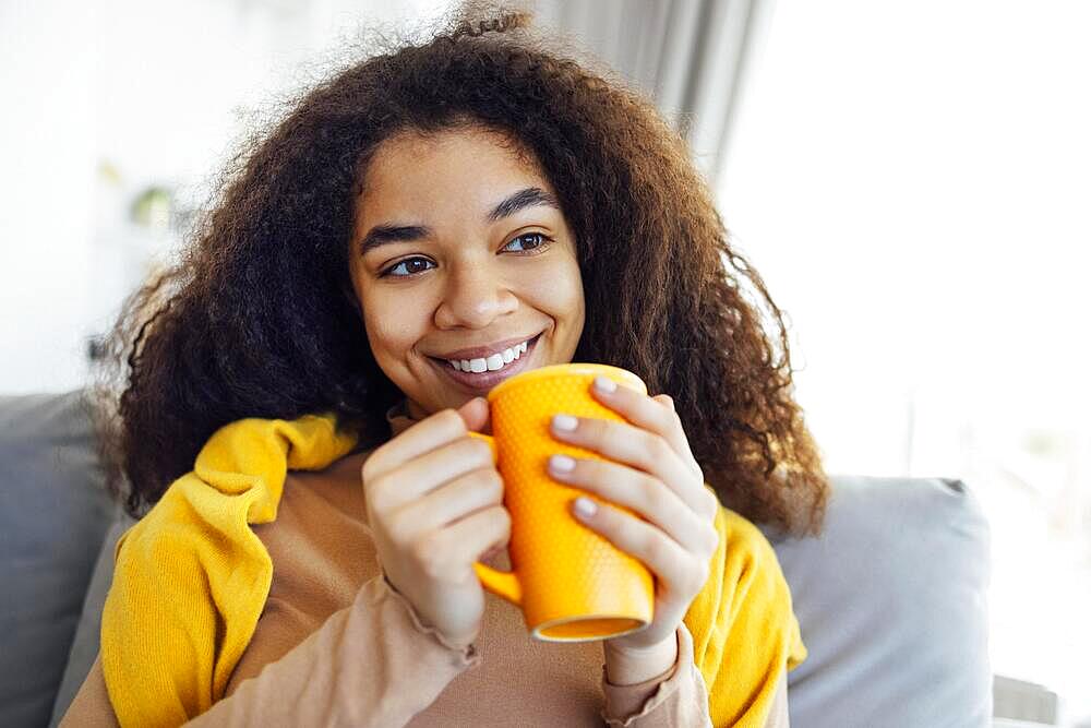 Cheerful smiling african american girl relaxing on couch with cup of coffee, resting at home, enjoying day off, wearing warm clothes