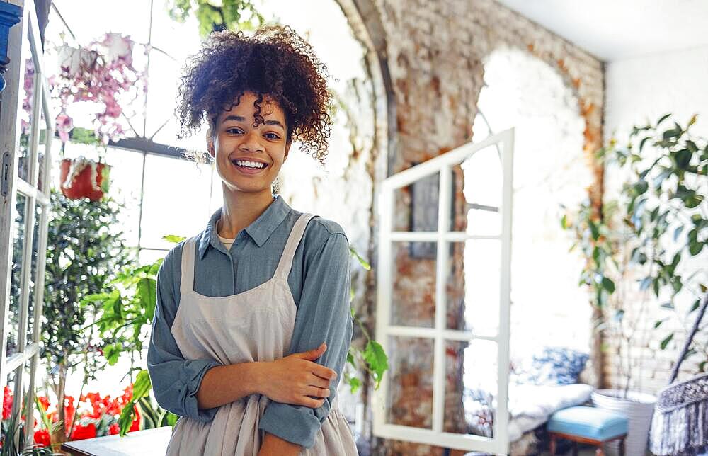 Young black woman wearing beige apron on flower shop background with copy space. Portrait of successful african american woman looking at camera