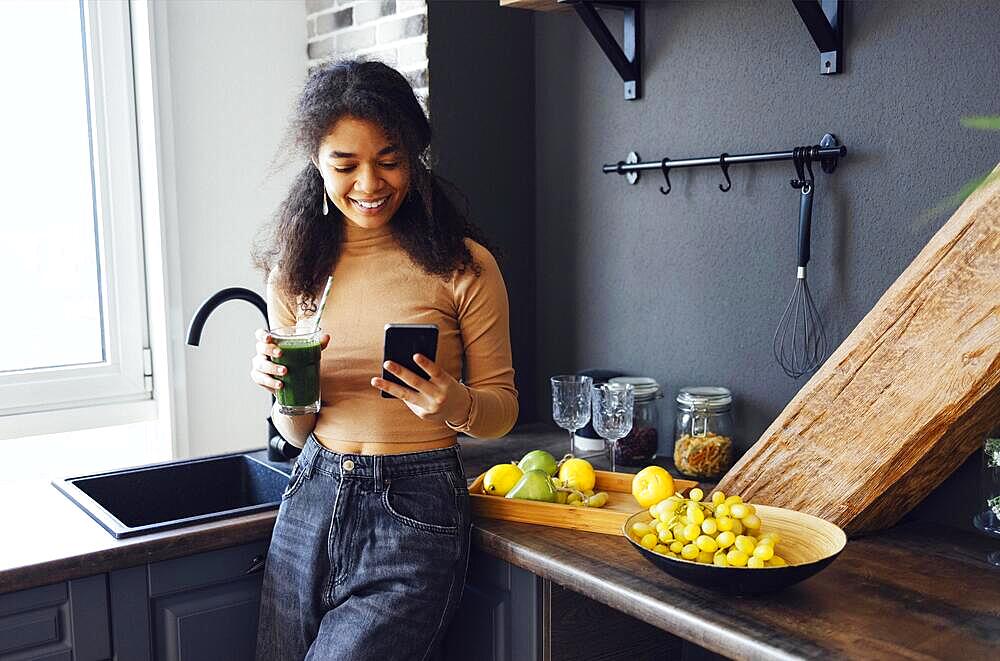 Afro woman holding a vegetables drink. Young african american female drinking green juice with reusable bamboo straw and reading news on mobile phone in loft apartment at the kitchen. Home concept. Healthy lifestyle concept. Copy space