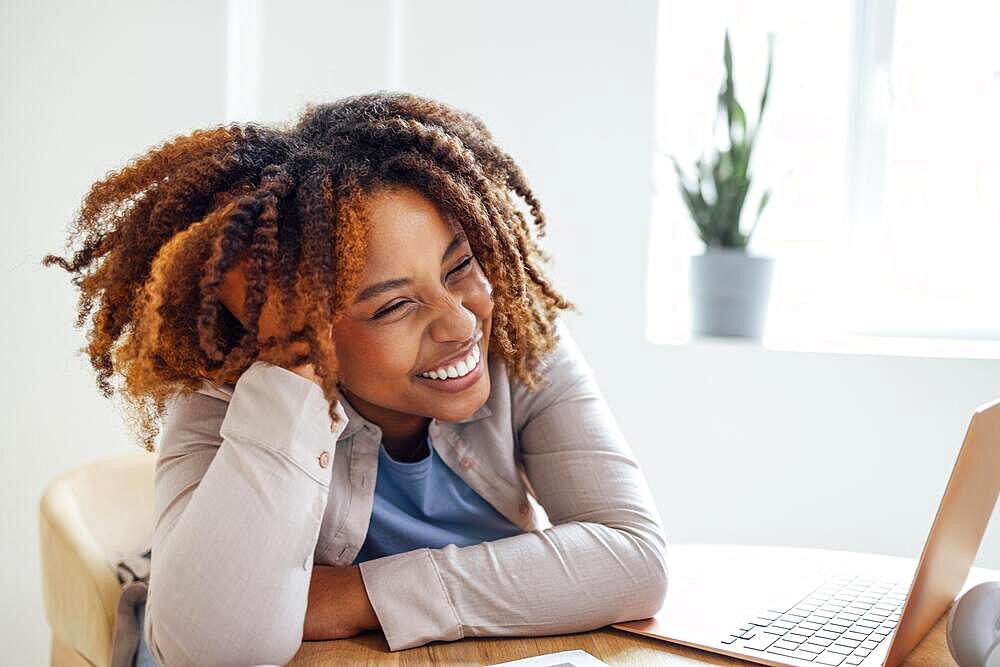 Young African American woman is working on a laptop. Focused female student is looking at computer screen and watching webinar or doing video chat by webcam. Remote education or home office concept