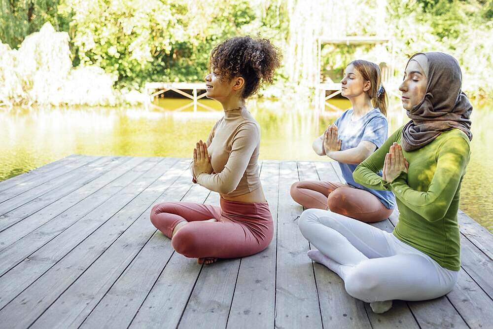 Group of teenages is engaged in fitness on pier by the lake or river. Young and positive girls go in for sports. Muslim in a hijab, african american and white females are doing stretching in the park