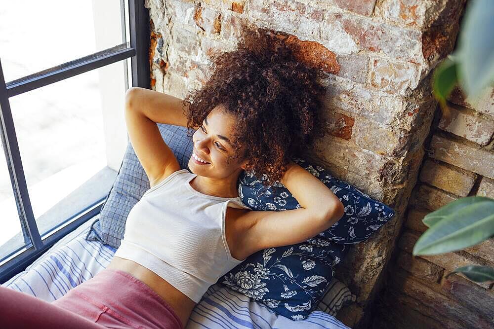 Pleased relaxed African American ethnicity girl in homewear sitting on windowsill, happy afro female at home smiling gently looking to the window