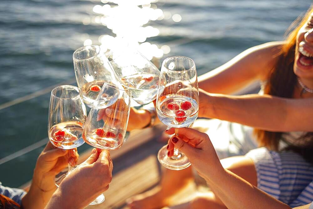 Female hands holding glasses of champagne with raspberries on board the boat. Sunset sky and sea on the background. Making a celebratory toast with sparkling wine. Birthday, holiday, party and friendship concept