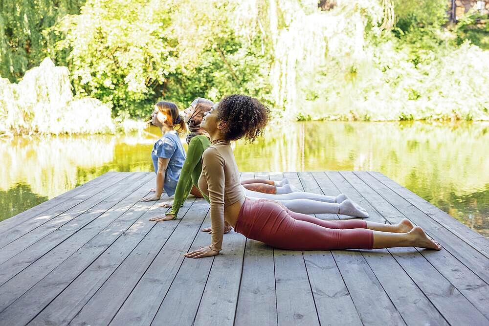 Group of teenages is engaged in fitness on pier by the lake or river. Young and positive girls go in for sports. Muslim in a hijab, african american and white females are doing stretching in the park