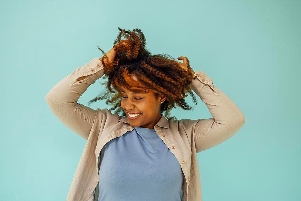 Positive african american female model smiling at camera and touching clean curly hair after hygiene treatment on blue background