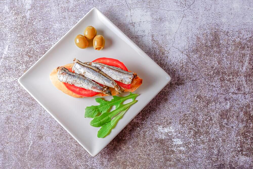 Tapa of sardines on a slice of bread with tomato and olives on a white plate with a typical spanish white background