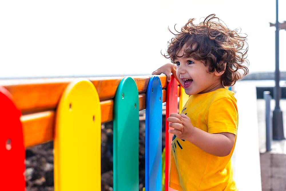 Cute little boy with big eyes and long hair smiling in a game together with the colored barriers of the park