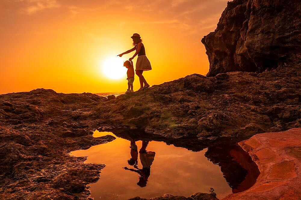 Silhouette of mother and son walking in the sunset on the beach of Tacoron in El Hierro, Canary Islands, vacation concept, orange sunset, walking by the sea pointing the path