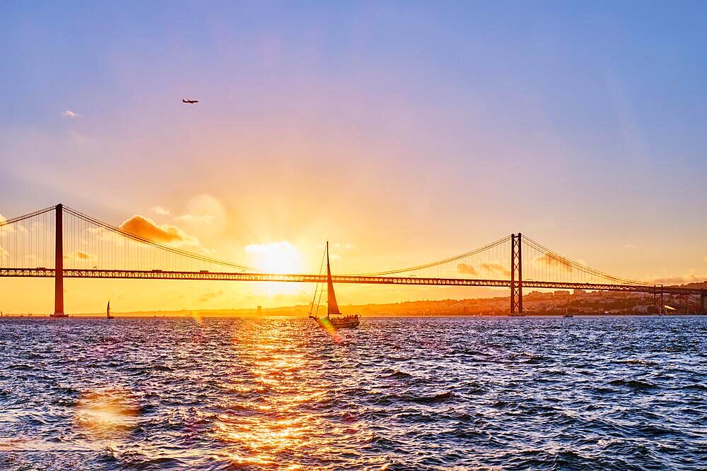 View of 25 de Abril Bridge famous tourist landmark of Lisbon connects Lisboa and Almada on Setubal Peninsula over Tagus river with tourist yacht silhouette at sunset and flying plane. Lisbon, Portugal, Europe