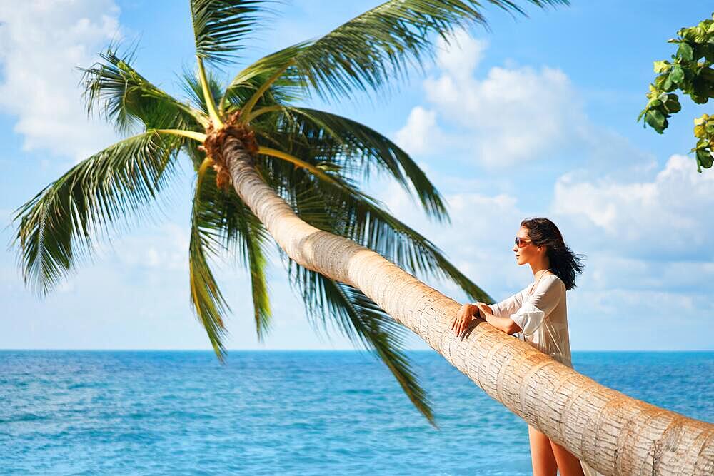 Pretty young woman in white bikini enjoy her tropical beach vacation. Beautiful woman rest under palm tree on the sea. Summer concept