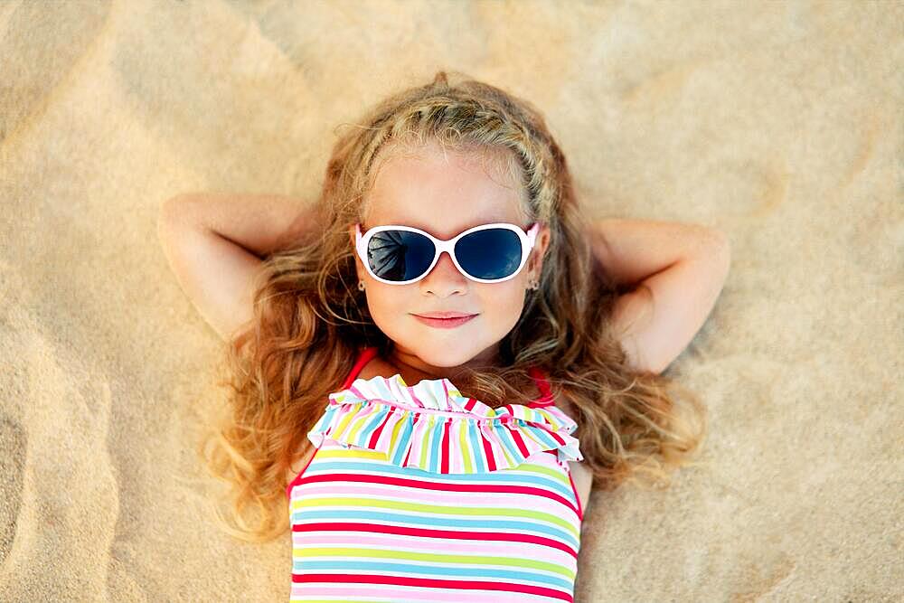 Top view of pretty little blonde girl in sunglasses lying on sandy beach during summer vacation. Sea relax, happy childhood concept