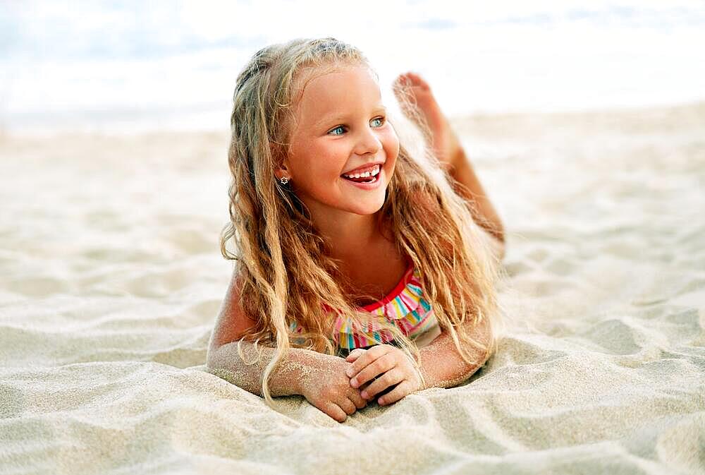 Adorable little blonde girl relax on sandy beach enjoy sea. Smiling pretty child posing on beach. Summer vacation, happy childhood and emotions concept