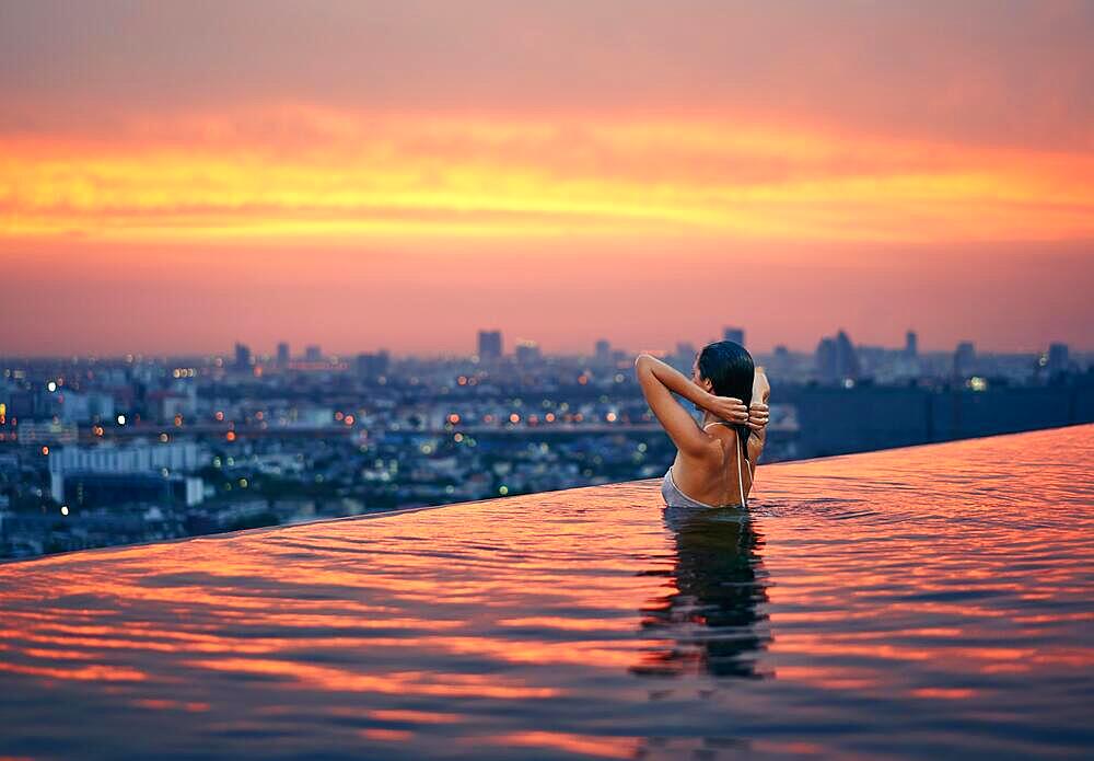 Young woman relax in swimming pool on roof top during amazing sunset and enjoy cityscape. Summer vacation and holidays concept