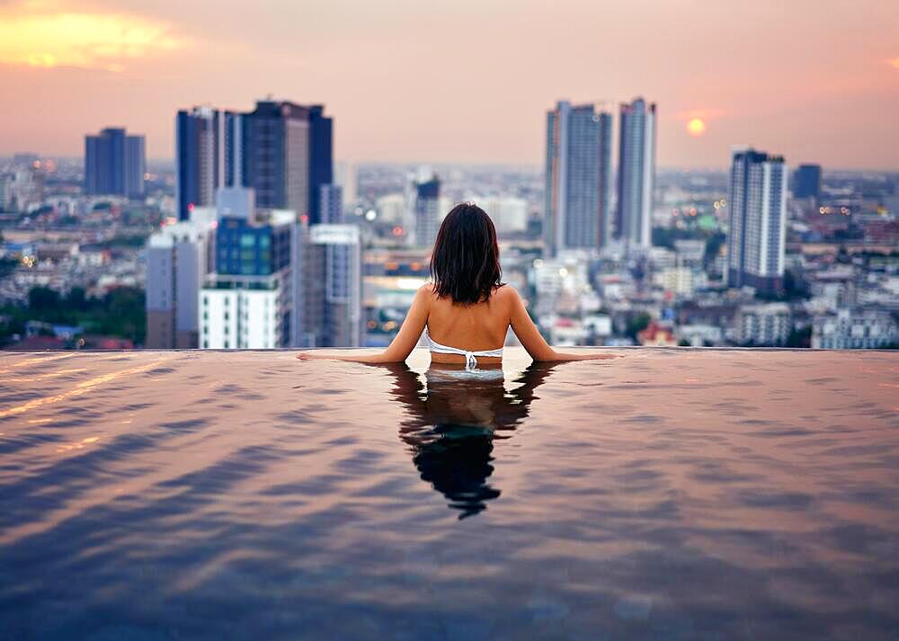 Young woman relax in swimming pool on roof top during amazing sunset and enjoy city view. Summer vacation and holidays concept