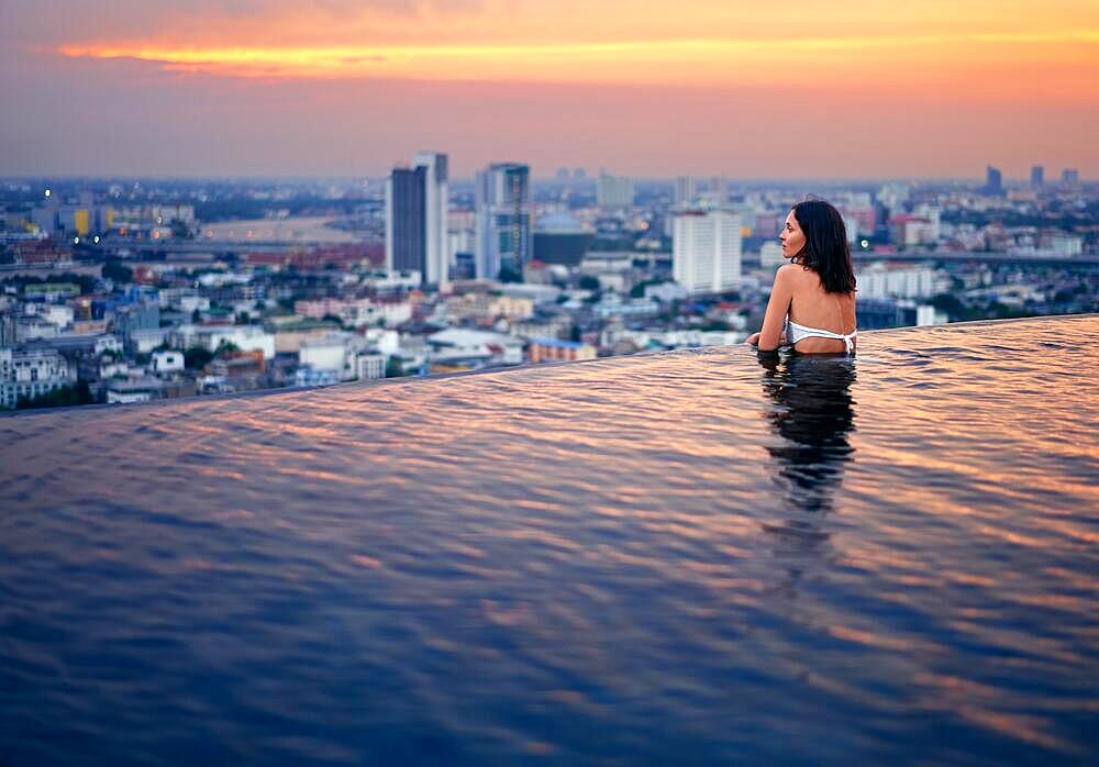 Young woman relax in swimming pool on roof top during amazing sunset and enjoy city view. Summer vacation and holidays concept