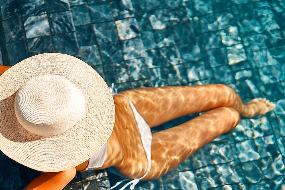 Unrecognizable woman in big hat with sexy slim body relaxing on the swimming pool. Top view